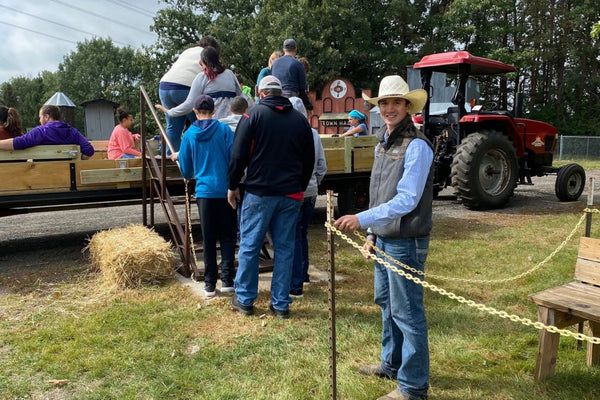 A group of people standing on a grassy field near a red tractor and a hay wagon at what appears to be a farm or rural event. A person in the foreground wears a cowboy hat, gray vest, blue shirt, and jeans while holding a rope barrier.