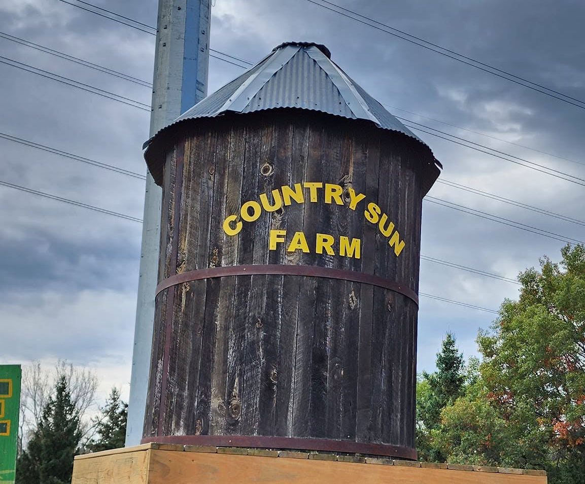 A rustic wooden water tower with a metal silo top and yellow lettering reading 'COUNTRY SUN FARM,' mounted on a wooden support structure outdoors under a cloudy sky.