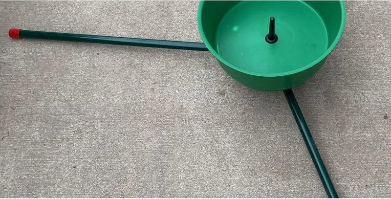 A green plastic Christmas tree stand with three legs and a red-tipped handle, placed on a gray concrete surface.
