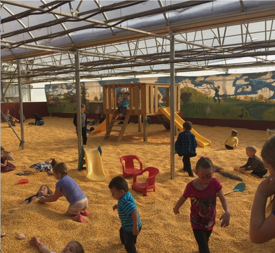 A children's corn pit playground with kids playing in a sandy play area under a translucent roof, featuring a wooden climbing structure, yellow slides, red plastic chairs, and painted mural walls in the background.