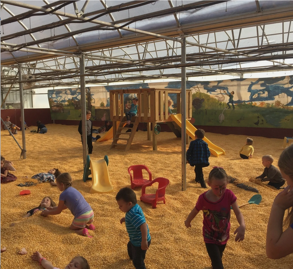 A children's corn pit playground with kids playing in a sandy play area under a translucent roof, featuring a wooden climbing structure, yellow slides, red plastic chairs, and painted mural walls in the background.