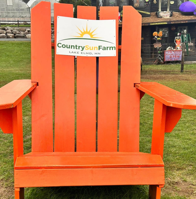 A bright orange Adirondack-style outdoor chair on grass, with a white sign reading Country Sun Farm and Lake Elmo, MN attached to the backrest. A farm setting with fencing and plants is visible in the background.